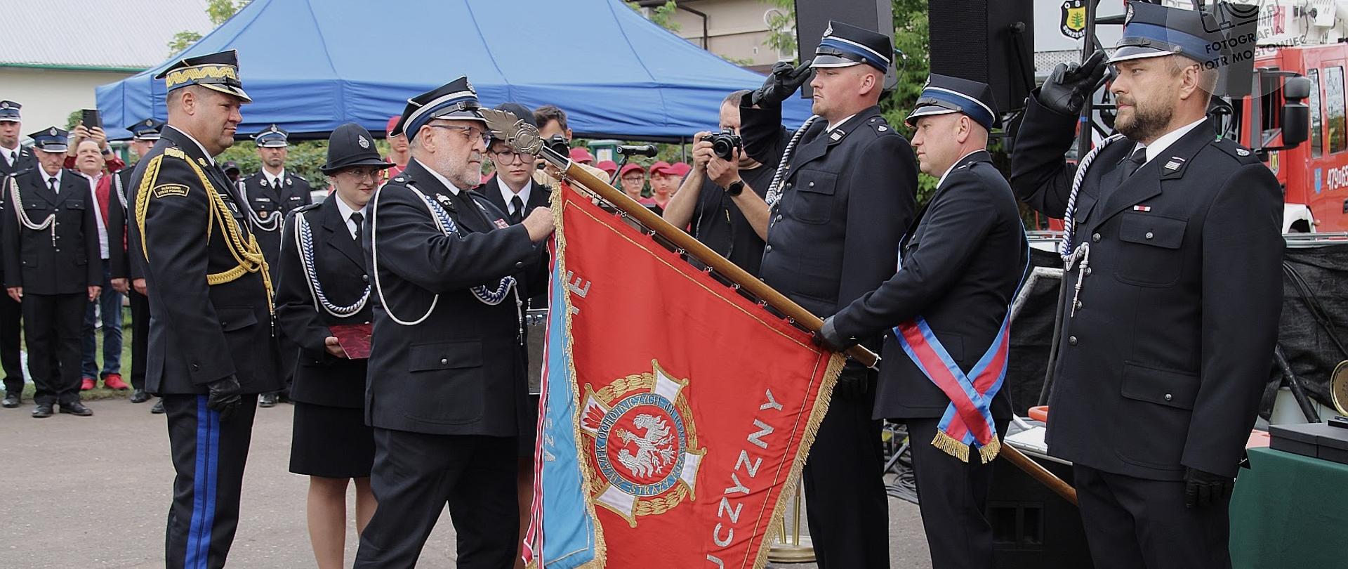 Na pierwszym planie kilku strażaków w galowych mundurach uczestniczy w ceremonii związanej ze sztandarem. Jeden ze strażaków trzyma czerwony sztandar, który jest pochylony w kierunku grupy. Obok stoją pozostali strażacy oraz osoby uczestniczące w uroczystości. W tle znajdują się namioty, drzewa i plac przed strażnicą. Jest jasno i pogodnie, a zdjęcie ma bardzo uroczysty i ceremonialny charakter.