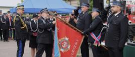 Na pierwszym planie kilku strażaków w galowych mundurach uczestniczy w ceremonii związanej ze sztandarem. Jeden ze strażaków trzyma czerwony sztandar, który jest pochylony w kierunku grupy. Obok stoją pozostali strażacy oraz osoby uczestniczące w uroczystości. W tle znajdują się namioty, drzewa i plac przed strażnicą. Jest jasno i pogodnie, a zdjęcie ma bardzo uroczysty i ceremonialny charakter.