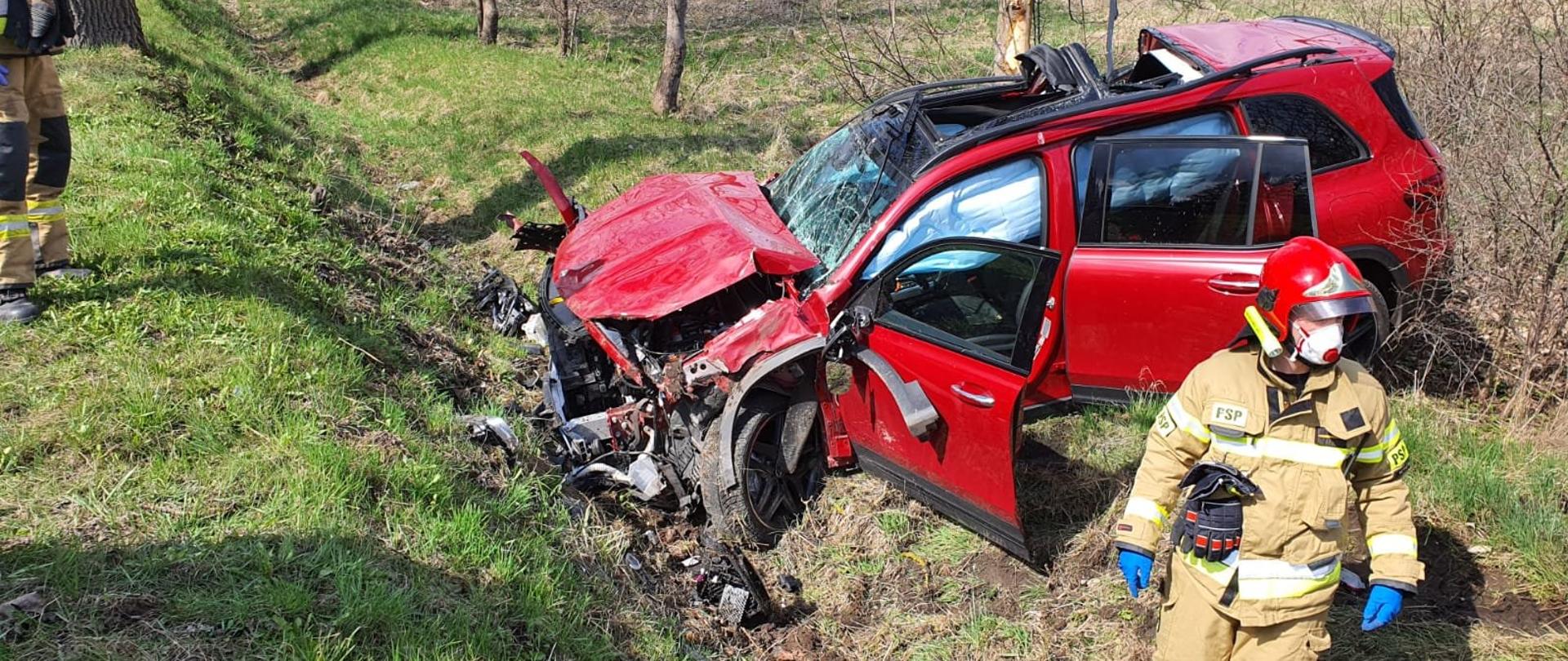 Zdjęcie przedstawia miejsce wypadku. Czerwony samochód całkowicie zniszczony znajduje się w rowie, dwóch strażaków w piaskowym ubraniu bojowym z założonym czerwonym hełmem oraz maseczkami ochronnymi znajdują się obok pojazdu.
