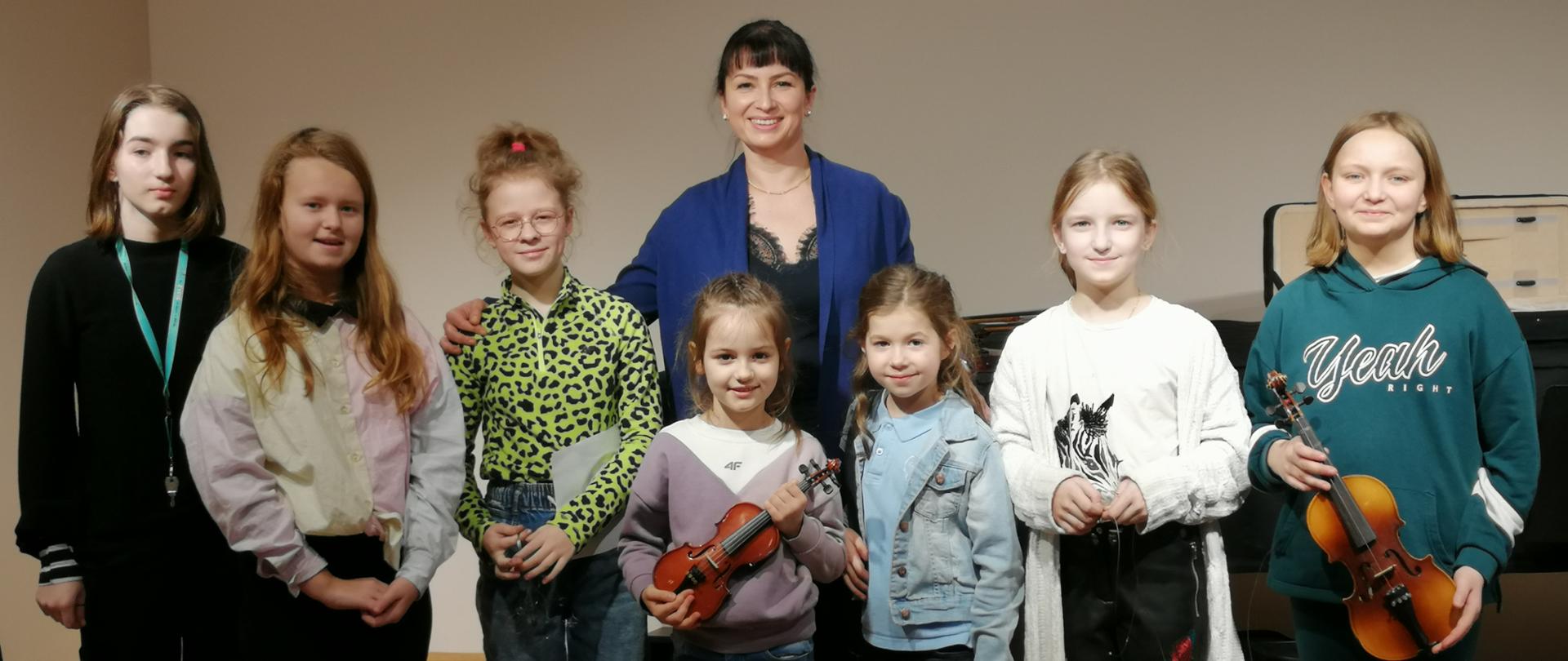 Zdjęcie przedstawia grupę uczniów wraz z nauczycielką prowadzącą warsztaty, dwie z uczennic trzymają w rękach skrzypce.