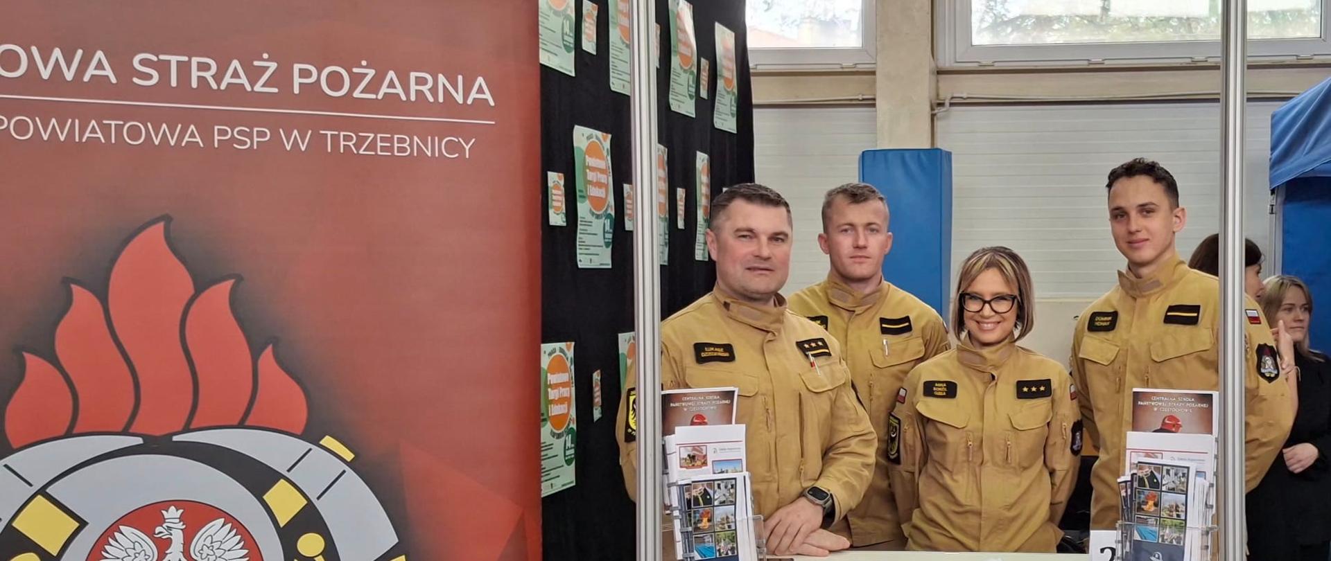 Zdjęcie przedstawia stoisko informacyjne Państwowej Straży Pożarnej podczas oficjalnego targów. Za białą ladą stoi czworo strażaków – trzech mężczyzn i jedna kobieta – ubranych w piaskowe mundury służbowe, którzy patrzą w stronę obiektywu. Nad nimi umieszczony jest biały szyld z wyraźnym napisem „Państwowa Straż Pożarna”. Po lewej stronie kadru znajduje się duży, bordowy baner reklamowy z widocznym, kolorowym godłem formacji przedstawiającym orła na tle hełmu strażackiego.