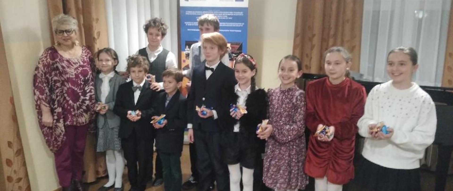 Zdjęcie przedstawia uczestników audycji fortepianowej wraz z nauczycielem. Dzieci elegancko ubrane trzymają w dłoniach cukierki.