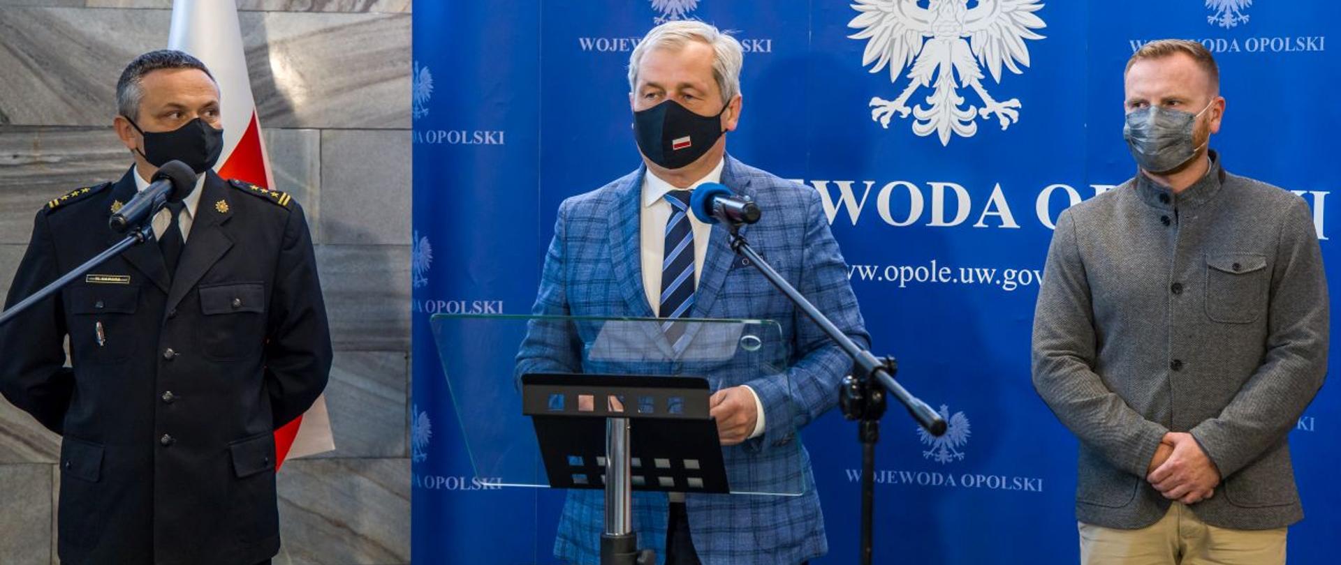 Wojewoda Opolski Sławomir Kłosowski na zdjęciu wraz z Zastępcą Opolskiego Komendanta Wojewódzkiego PSP w Opolu st. bryg. Wojciechem Nawarą oraz Kierownikiem CPR Ditmarem Kurzałem podczas konferencji w siedzibie Opolskiego Urzędu Wojewódzkiego. W tle niebieski banner z białymi napisami Wojewoda Opolski.