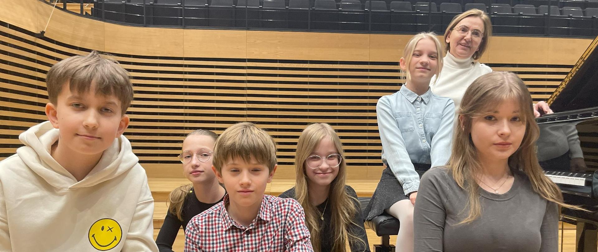 Cztery dziewczynki i dwóch chłopców siedzą lub kucają przy fortepianie, za nimi stoi kobieta.