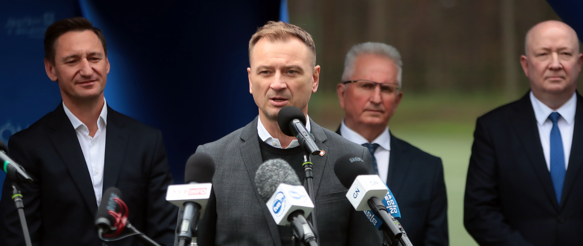 Zdjęcie przedstawia Ministra Sportu i Turystyki Sławomira Nitrasa przemawiającego do mikrofonu w czasie konferencji prasowej. Pan minister jest ubrany w białą koszulę, sweter oraz szarą marynarkę, w tle stoją trzej mężczyźni ubrani w garnitury, a za nimi jest kawałek niebieskiego namiotu i naturalna zieleń.