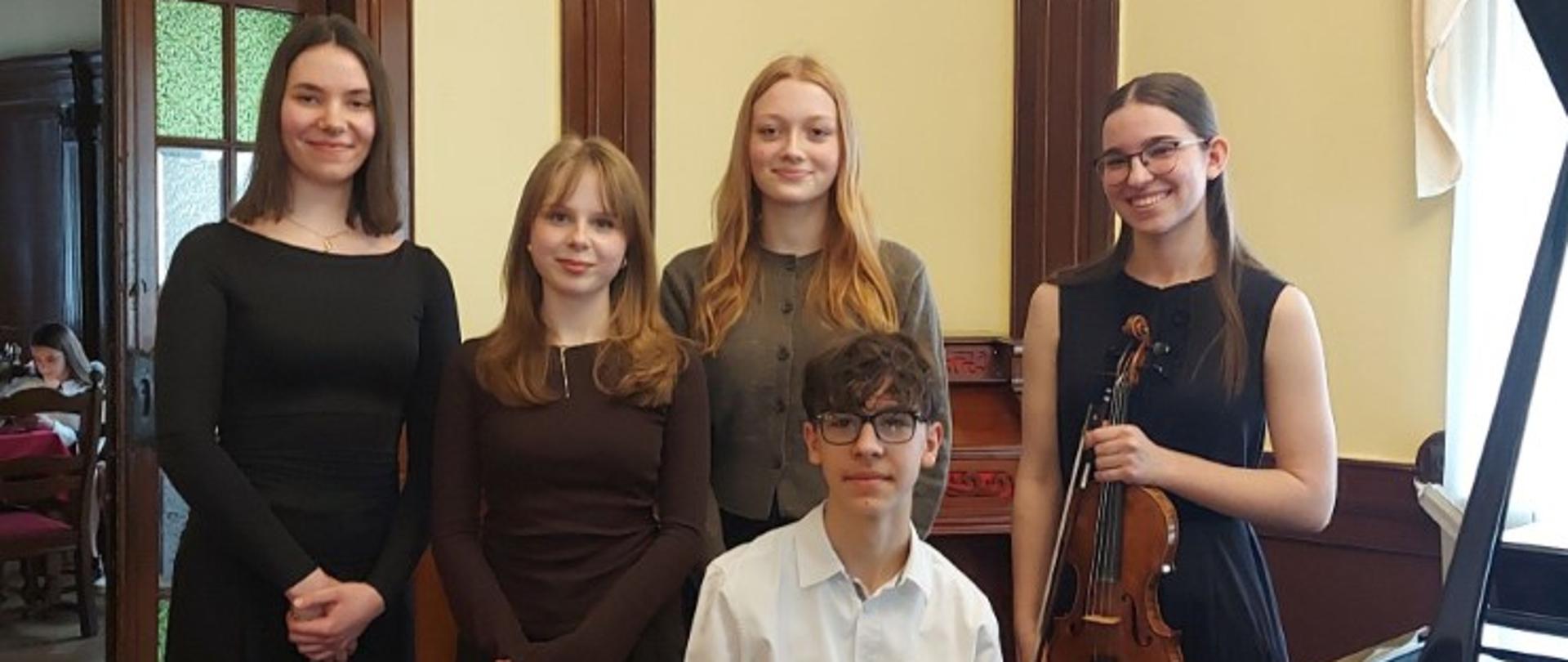 Zdjęcie kolorowe. Cztery młode dziewczyny i młody chłopiec w eleganckich strojach. Po lewej dziewczyna w czarnym dopasowanym komplecie z długimi rękawami i szerokimi spodniami. Obok niej niższa dziewczyna w brązowej bluzce z długim rękawem i ciemnych spodniach. W środku, z tyłu, dziewczyna w szarym swetrze. Przed nią siedzący przy czarnym fortepianie chłopiec w białej koszuli i okularach. Po prawej dziewczyna w czarnej sukience bez rękawów, trzymająca skrzypce i smyczek. Grupa pozuje w klasycznym wnętrzu z drewnianymi zdobieniami na ścianach i wzorzystym dywanem. W tle, przez otwarte drzwi, fragment innego pomieszczenia ze stolikiem.