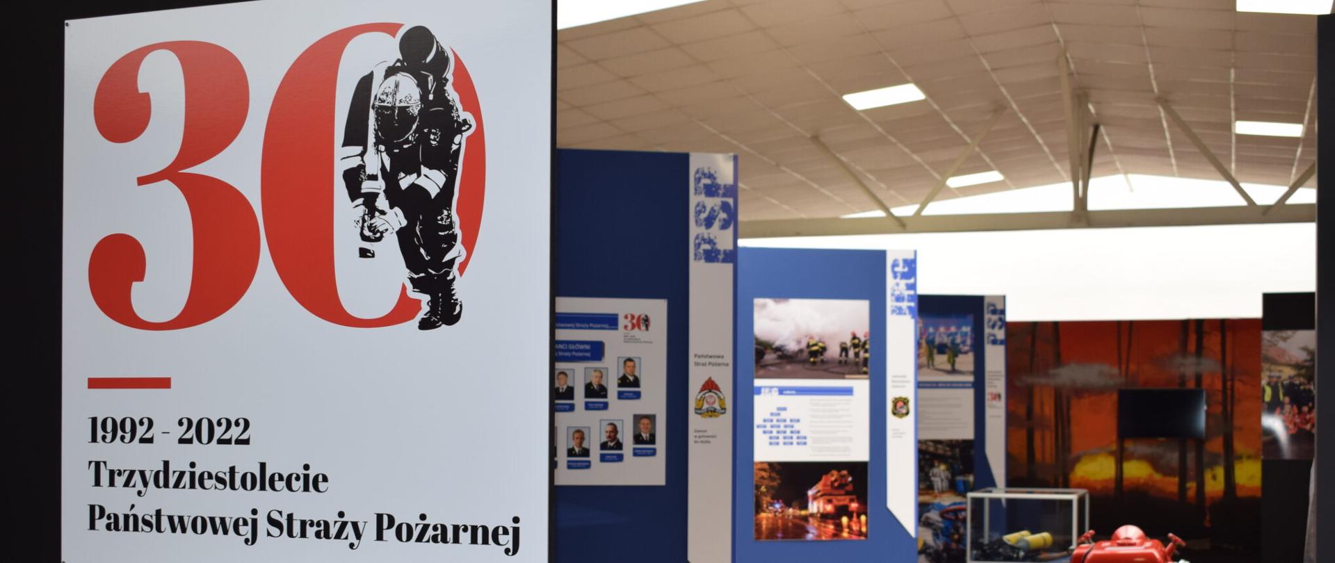 Wnętrze sali wystawowej w muzeum, na pierwszym planie plakat z logotypem 30 lecia PSP, w dalszym planie plakaty z: zdjęciami komendantów, strażacy w akcji, pożar i inne, na środku stoi motopompa
