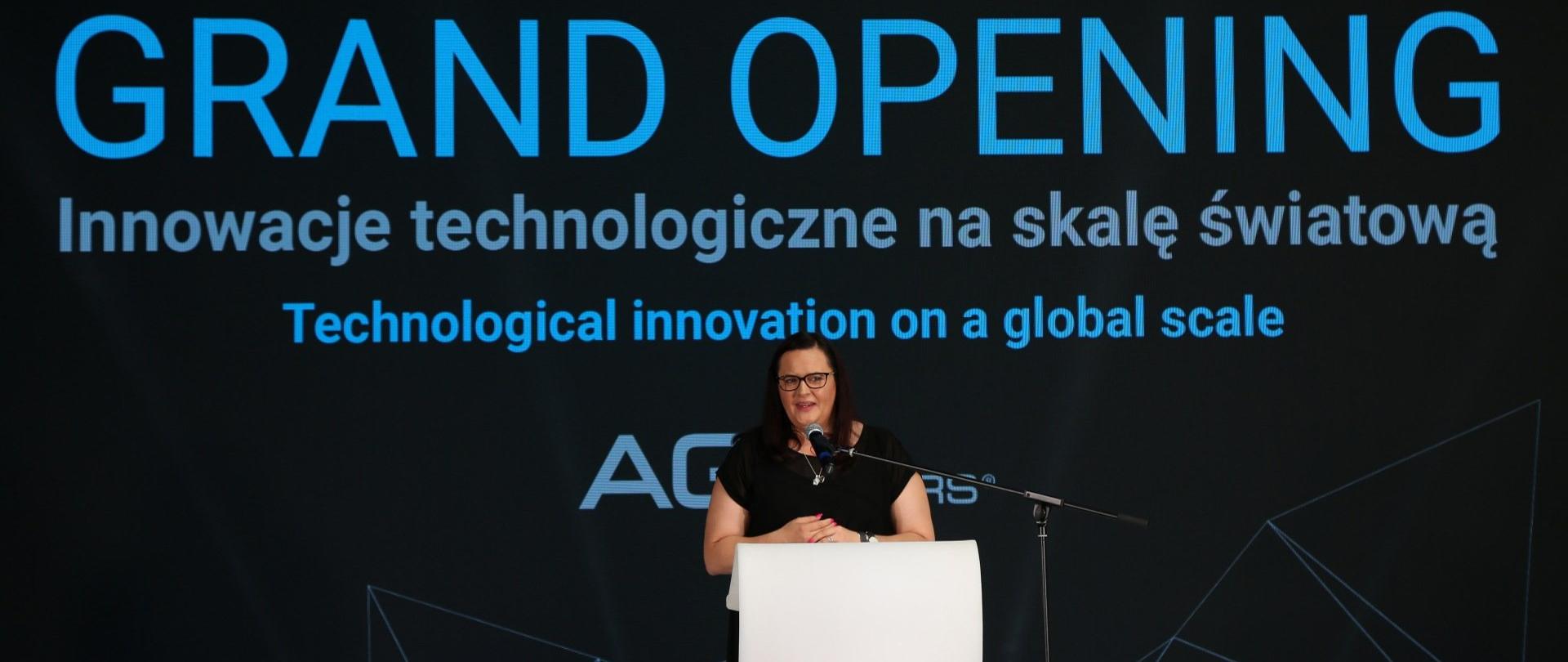 Wiceminister Małgorzata Jarosińska-Jedynak stoi w białej mównicy i przemawia do mikrofonu. Za nią na ekranie na czarnym tle wyświetla się napis: GRAND OPENING Innowacje technologiczne na skalę światową"