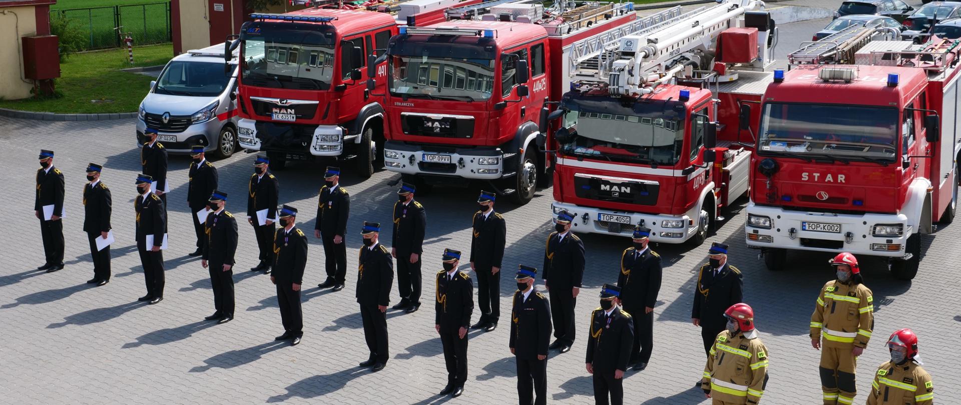 Zdjęcie przedstawia strażaków KP PSP w Opatowie stojących w dwuszeregu, samochody strażackie oraz budynki komendy. 