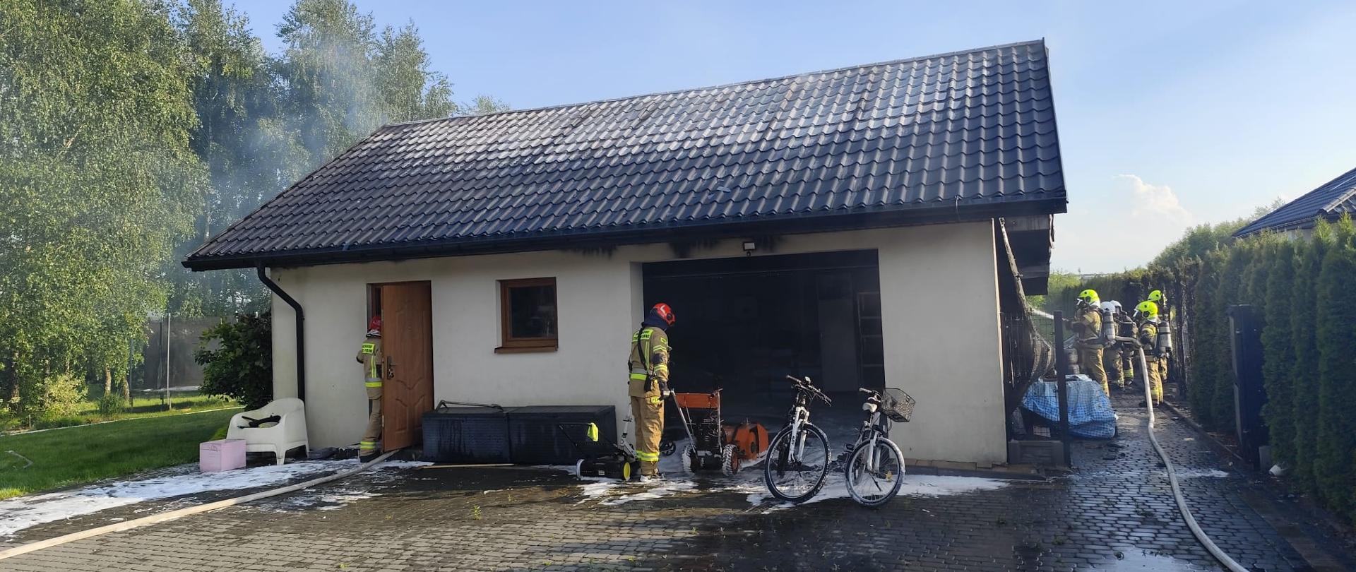 Zdjęcie przedstawia front budynku garażowego z otwartymi wrotami. Po prawej stronie widać pracujących strażaków.