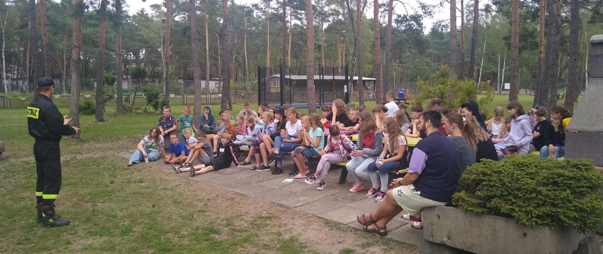 po lewej stronie stoi strażak na przeciwko niego siedzi grupa dzieci około 40 osób. Zdjęcie wykonane na zewnątrz 
