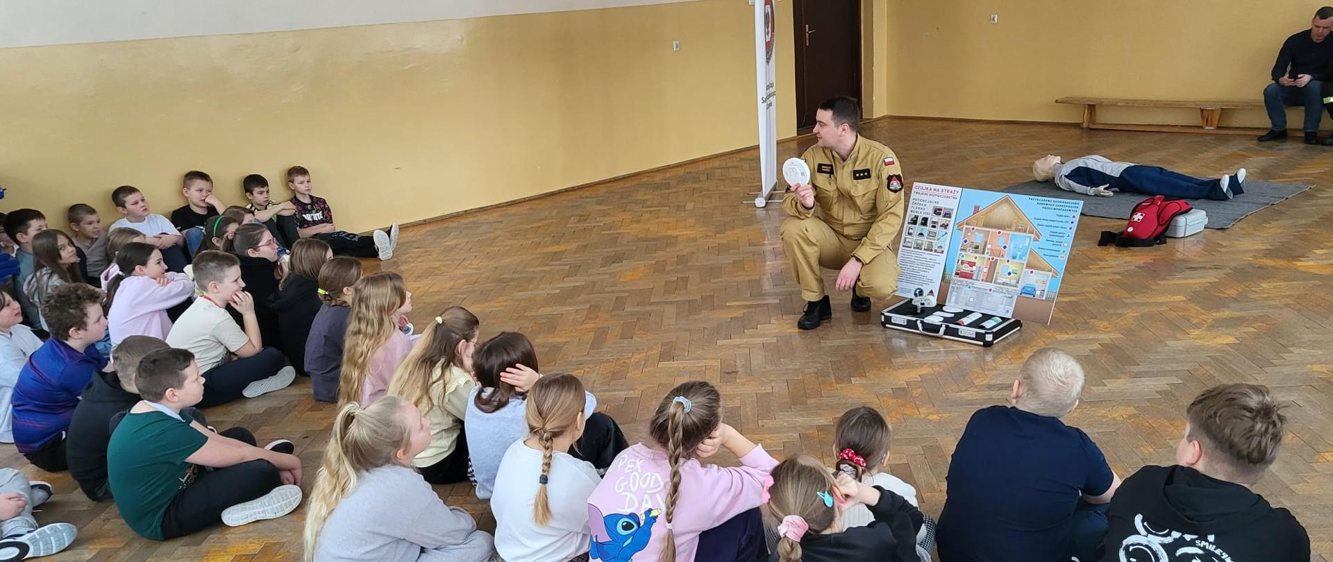 Strażak w piaskowym mundurze prowadzi zajęcia edukacyjne dla grupy dzieci siedzących na podłodze w sali gimnastycznej. Podczas spotkania funkcjonariusz prezentuje czujkę dymu oraz plansze instruktażowe dotyczące bezpieczeństwa przeciwpożarowego i pierwszej pomocy.
