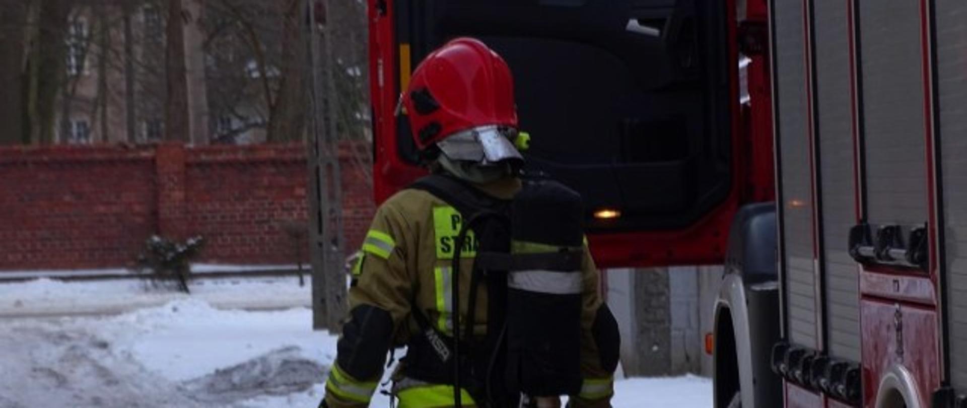 Po prawej samochód pożarniczy, strażak odwrócony tyłem w aparacie powietrznym.