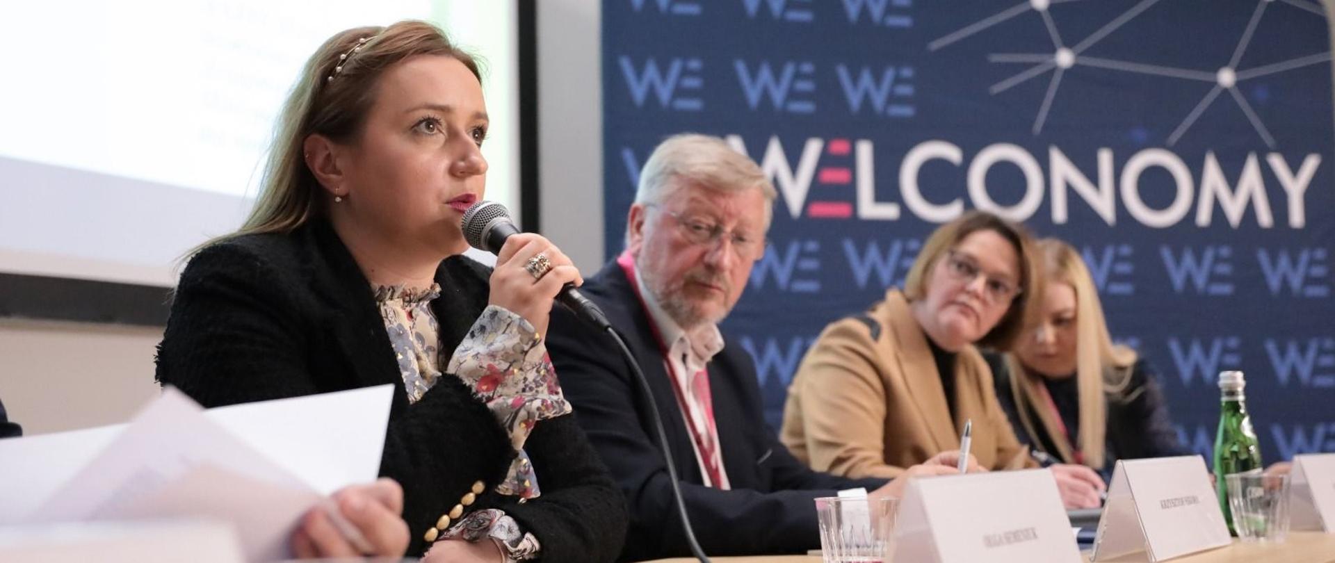 Wiceminister Olga Semeniuk podczas Welconomy Forum in Toruń. Na zdjęciu widać minister, która zabiera głos i pozostałych panelistów. Uczestniczy siedzą przy stole.