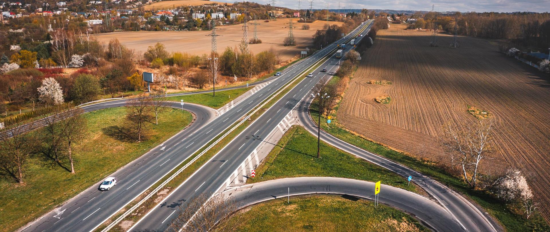 Zdjęcie lotnicze przedstawiające skrzyżowanie i fragment dwujezdniowej drogi krajowej biegnącej przez rolniczy, pagórkowaty krajobraz. Widoczna jest estakada oraz rondo pod nią, a także pola uprawne, pojedyncze drzewa i zabudowania w tle. Na niebie znajdują się rozproszone chmury. W prawym dolnym rogu umieszczono podpis: FOT. PAWEŁ KLARECKI/GDDKiA