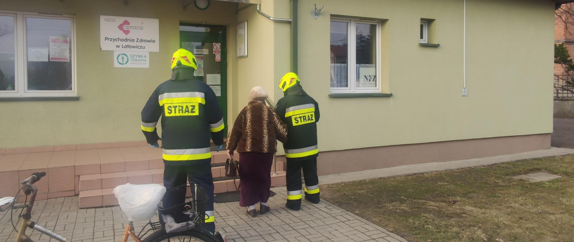 Strażacy podprowadzają starszą kobietę na szczepienie do przychodni
