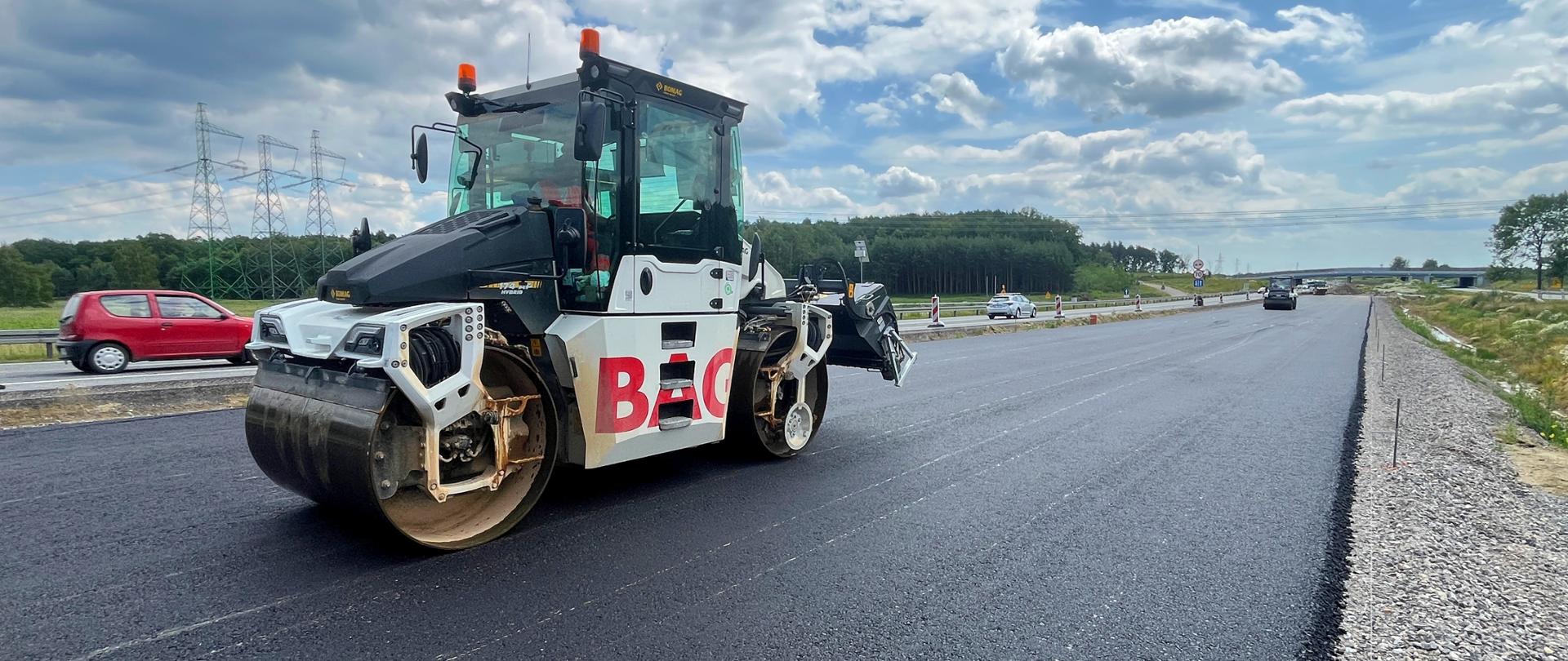 Widok z poziomu ziemi walec. Widać wykonywanie warstwy podbudowy. W tle pracują inne walce i maszyny budowlane.