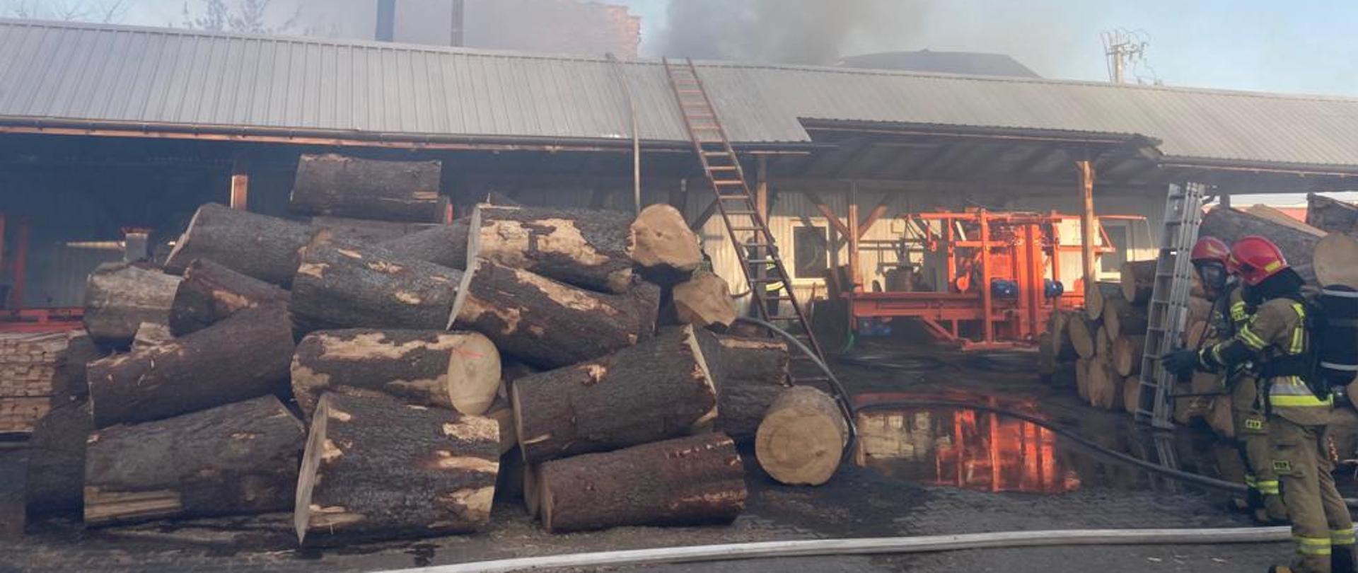 Zdjęcie przedstawia akcję ratowniczo - gaśniczą podczas pożaru hali produkcyjnej w Kuźnicy Grabowskiej. Na zdjęciu widzimy strażaków ubranych w aparatu ochrony układu oddechowego biorących bezpośredni udział w działaniach.Widoczny jest także palący się budynek hali oraz składowisko drewna Zdjęcie wykonane w porze dziennej na terenie zakładu.