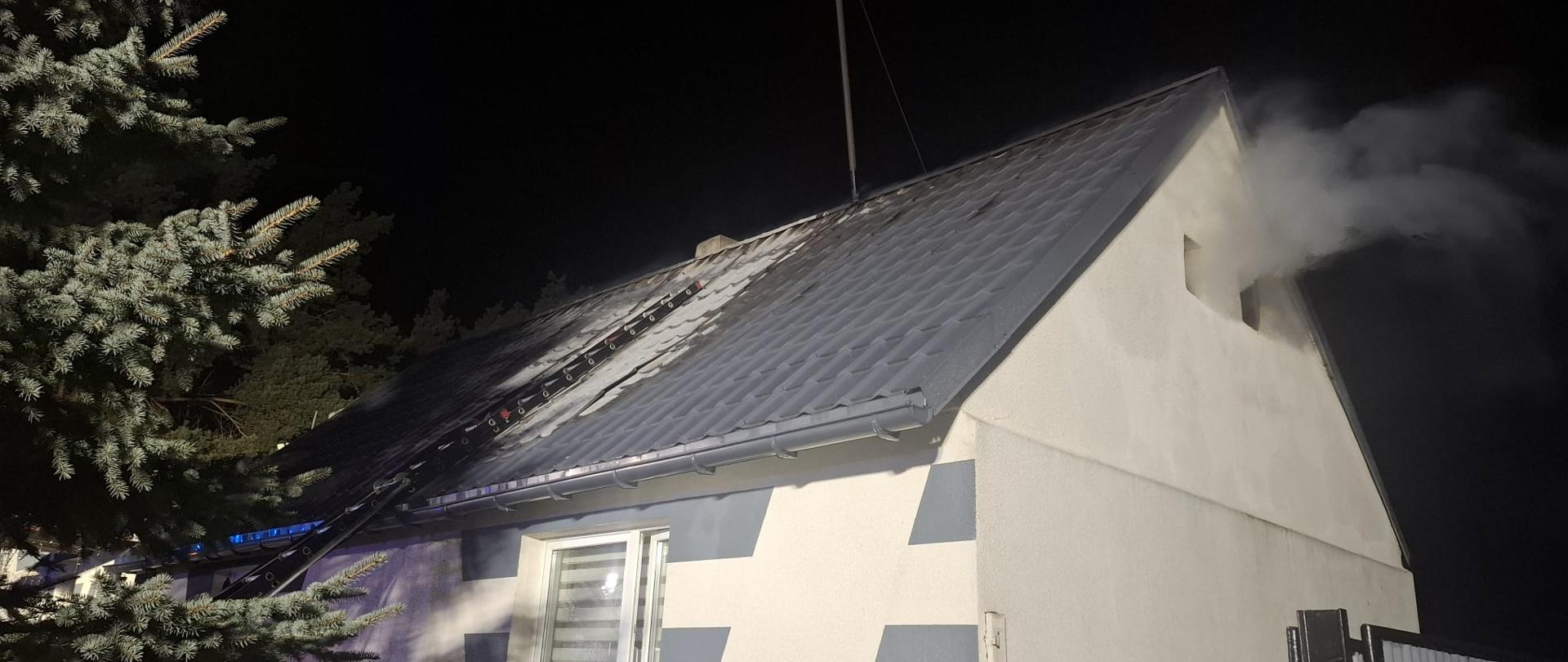 Zdjęcie przedstawia strażaków podczas gaszenia pożaru dachu budynku.
W tle budynek.
