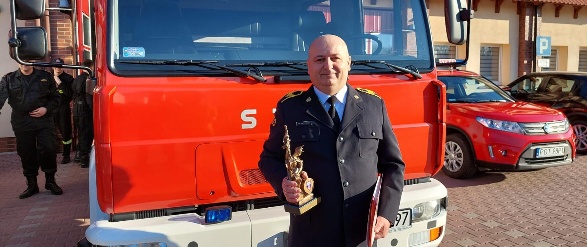 Zdjęcie przedstawia uroczystą zmianę służby podczas której na zaopatrzenie emerytalnie przeszedł st. ogn. Grzegorz Matysiak. Na zdjęciu widzimy st. ogn. Grzegorza Matysiaka na tle samochodu pożarniczego. Zdjęcie wykonane w porze dziennej przed garaże JRG.