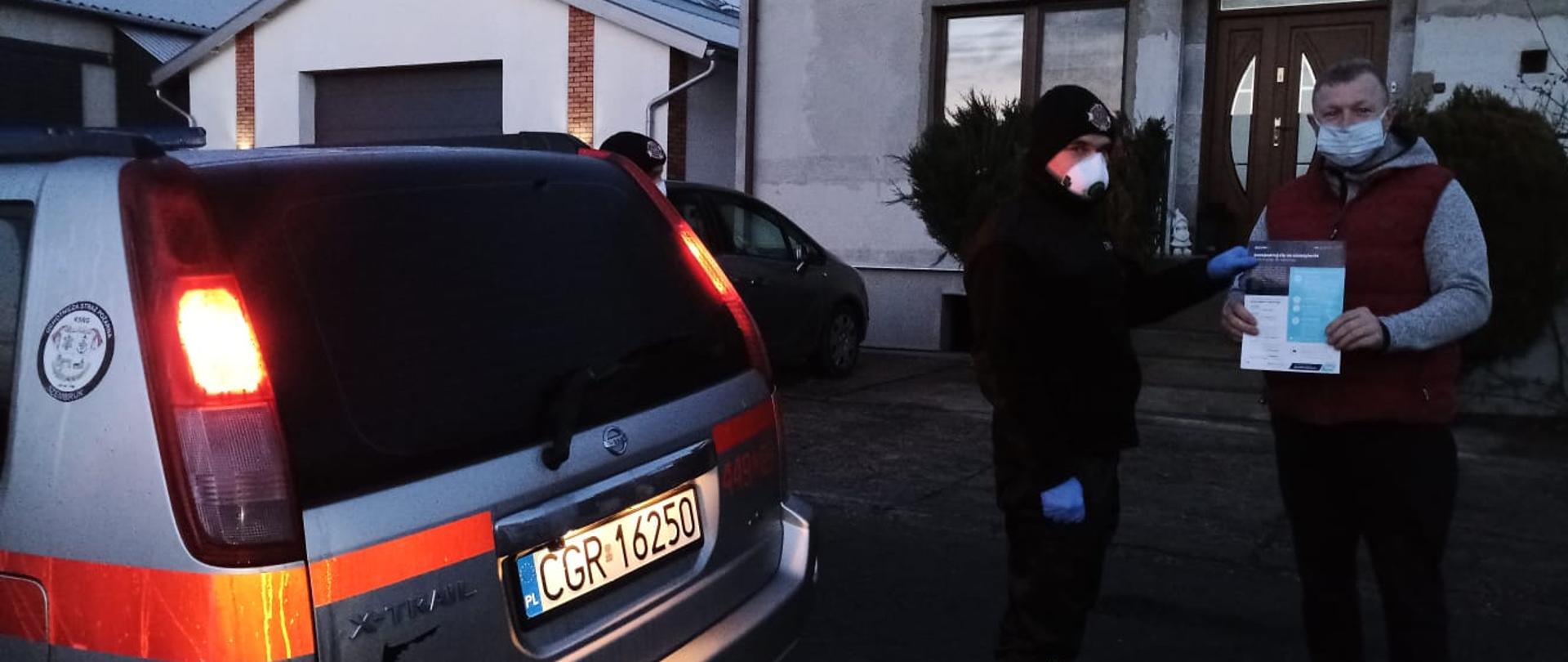 Zdjęcie przedstawia strażaka w ubraniu koszarowym przekazującego ulotkę informacyjną mężczyźnie. Obaj mają założone maseczki ochronne. Po lewej stronie widoczna tylna cześć srebrnego pojazdu strażackiego. W tle fasady domu mieszkalnego i budynków gospodarczych
