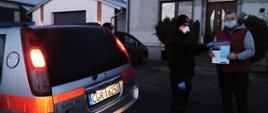 Zdjęcie przedstawia strażaka w ubraniu koszarowym przekazującego ulotkę informacyjną mężczyźnie. Obaj mają założone maseczki ochronne. Po lewej stronie widoczna tylna cześć srebrnego pojazdu strażackiego. W tle fasady domu mieszkalnego i budynków gospodarczych