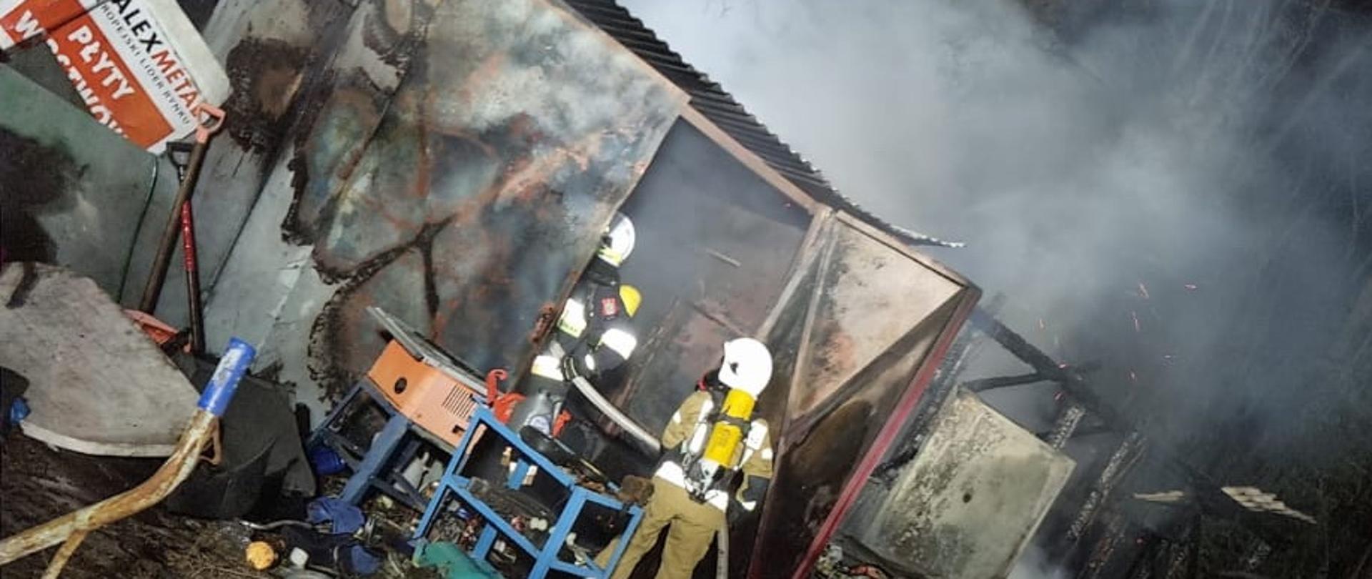 Dwóch strażaków zabezpieczonych aparatami ochrony dróg oddechowych wprowadza linię gaśniczą do zniszczonego przez pożar metalowego garażu, na pierwszym planie widoczne wyniesione z budynku narzędzia