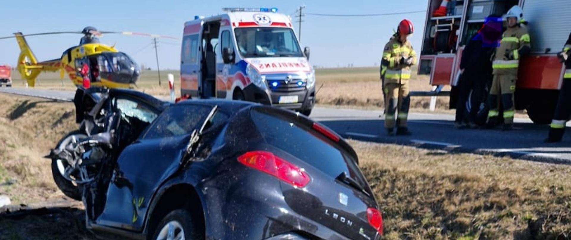 Na zdjęciu znajduje się wrak pojazdu na tle pola, strażacy, śmigłowiec LPR, karetka pogotowie i wóz straży pożarnej