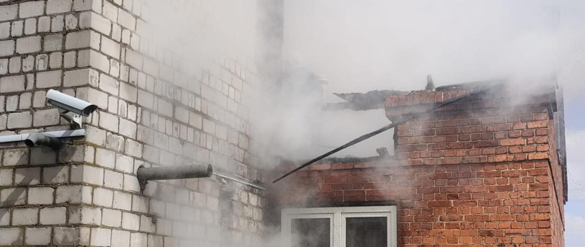 Na zdjęciu widać budynek po pożarze. Z części dachu wydobywa się dym. Ściany budynku są z cegieł, a na ziemi leżą zniszczone fragmenty okien i inne resztki.