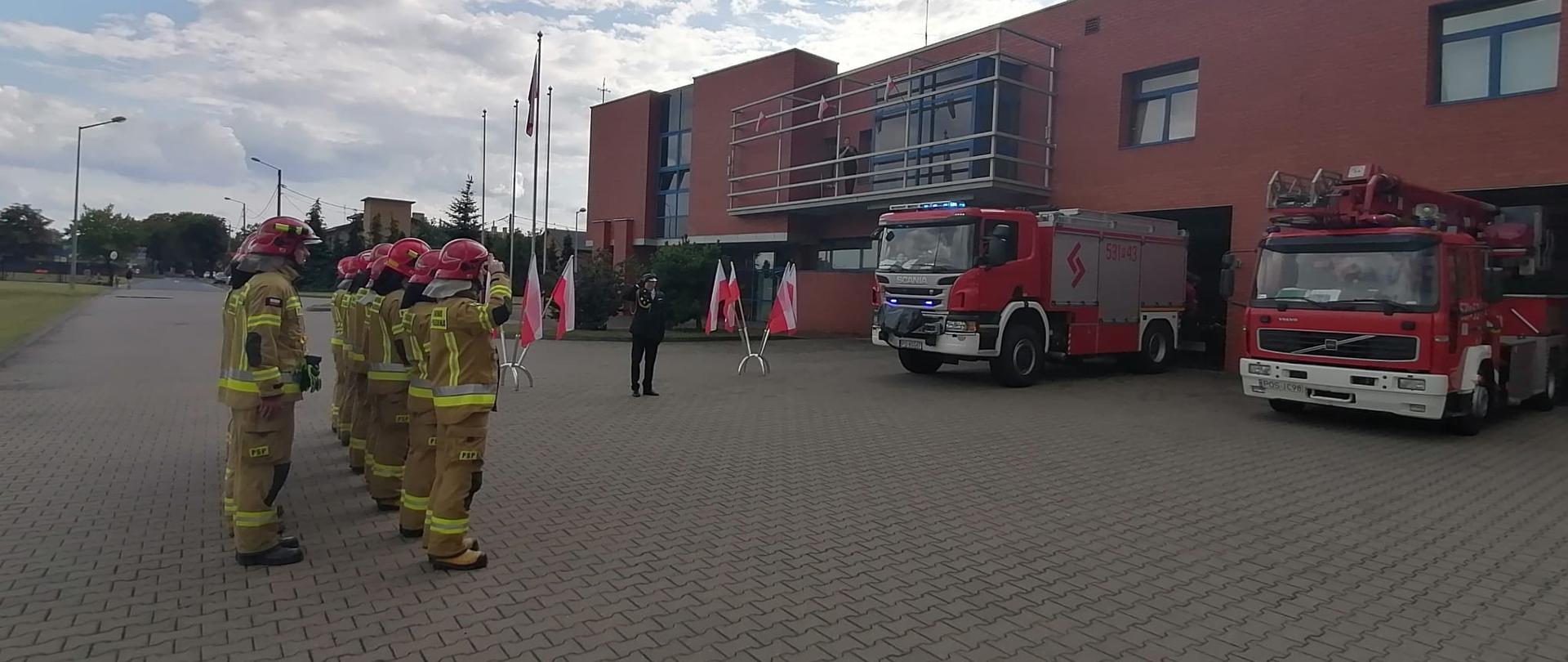 Strażacy w dwuszeregu w ubraniach specjalnych oddają hołd Powstańcom Warszawskim wspólnie z galowo umundurowanym Komendantem. W tle strażnica oraz cztery pojazdy gaśnicze.
