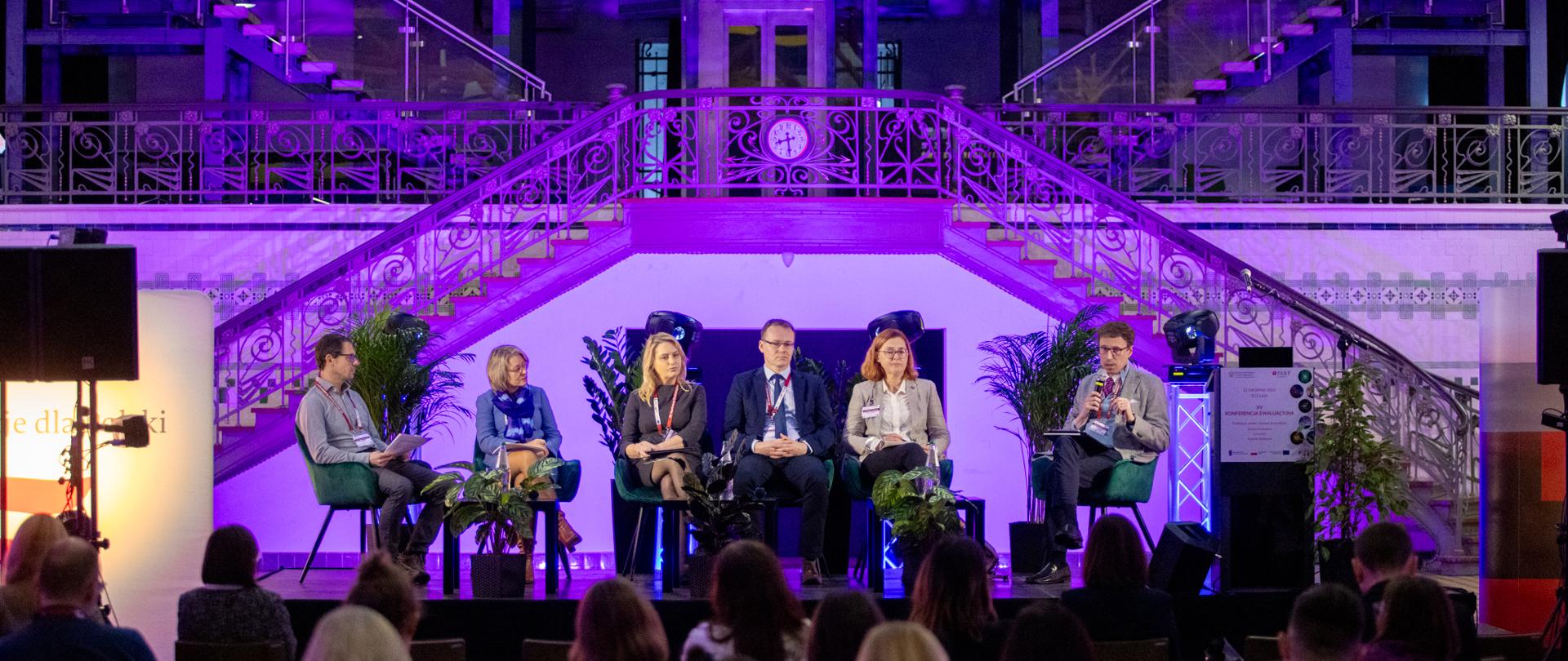 Kilka osób siedzi na krzesłach na scenie i rozmawiają 