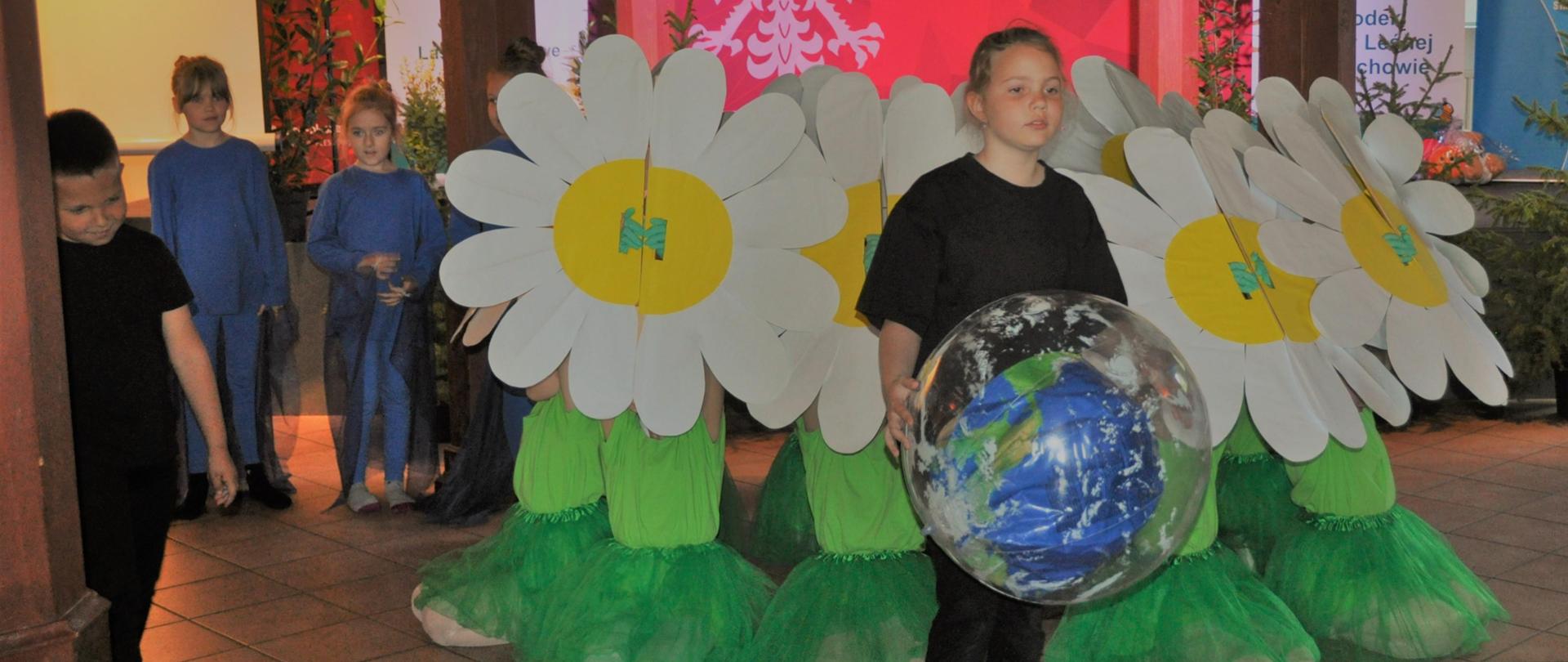 dziewczynka stoi na scenie z dużą piłką - ziemią. w tle dzieci kwaitki