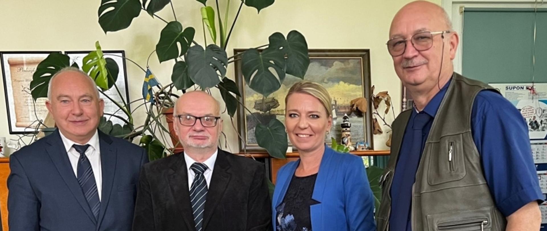 Grupa czterech osób w pokoju; w tle rośliny doniczkowe, obrazy i dokumenty.