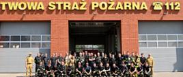 grupa strażaków po zakończonym egzaminie na tle budynku Komendy Powiatowej Straży Pożarnej z czerwonej cegły, jedna z siwych bram otwarta