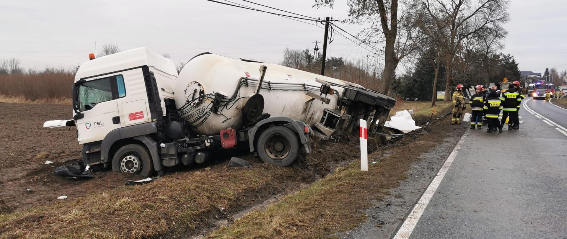 Przedstawia uszkodzony ciągnik siodłowy wraz z naczepą. Zestaw znajduję się poza pasem drogowym przewrócony na bok. Samochód i cysterna są mocno uszkodzone. Na pasie drogowym widać strażaków podczas akcji