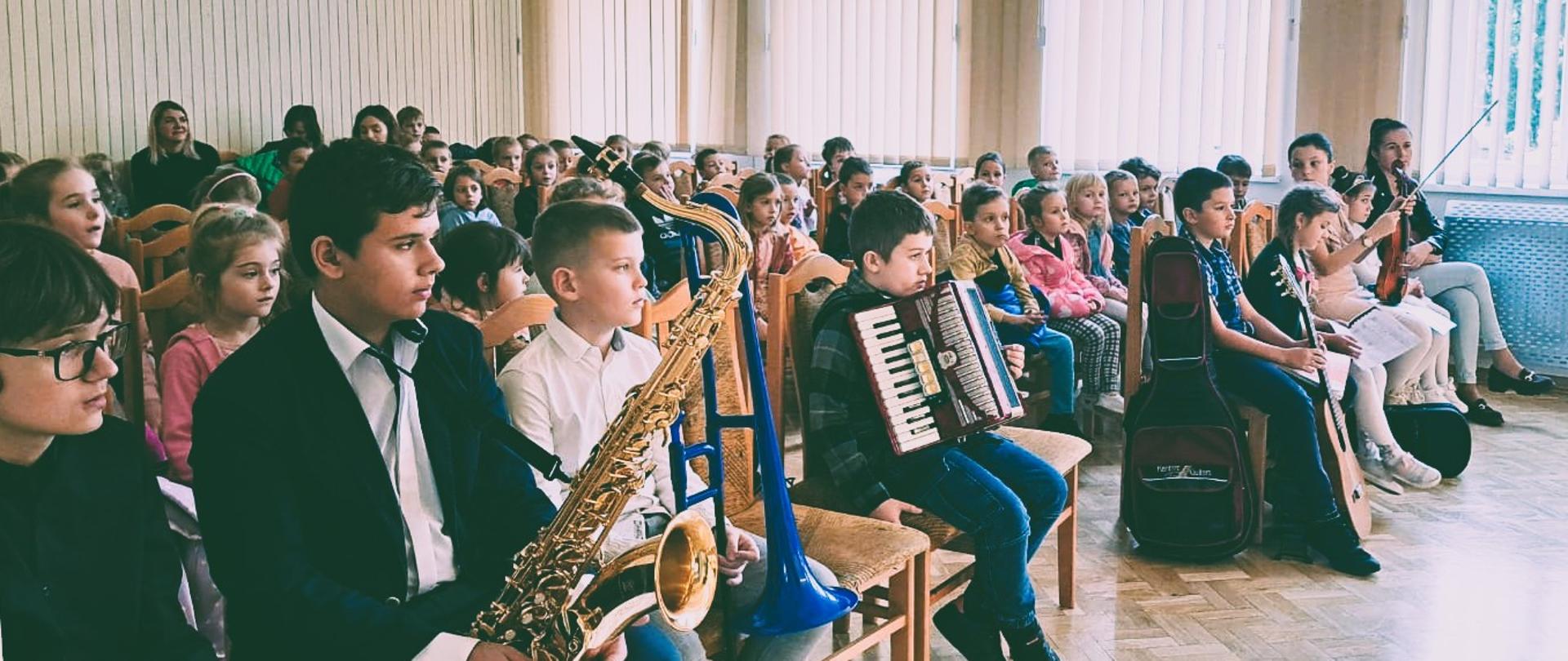 Zdjęcie przedstawia grupę dzieci siedzących w szkolnej auli z instrumentami muzycznymi. Aula ma duże okno z zasłonami i rośliną w rogu. Dzieci na drewnianych krzesłach trzymając instrumenty saksofon akordeon i skrzypce.