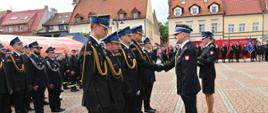Na pierwszym planie oficer w ciemnym mundurze galowym podaje rękę jednemu ze strażaków stojących w rzędzie na baczność. Obok nich stoi strażaczka w mundurze wyjściowym, która asystuje przy ceremonii. Wszyscy funkcjonariusze ubrani są w galowe mundury z czapkami rogatywkami i złotymi sznurami. W tle widoczny jest długi szereg innych strażaków, poczty sztandarowe przy czerwonej ściance promocyjnej oraz zabytkowe kamienice o jasnych elewacjach.