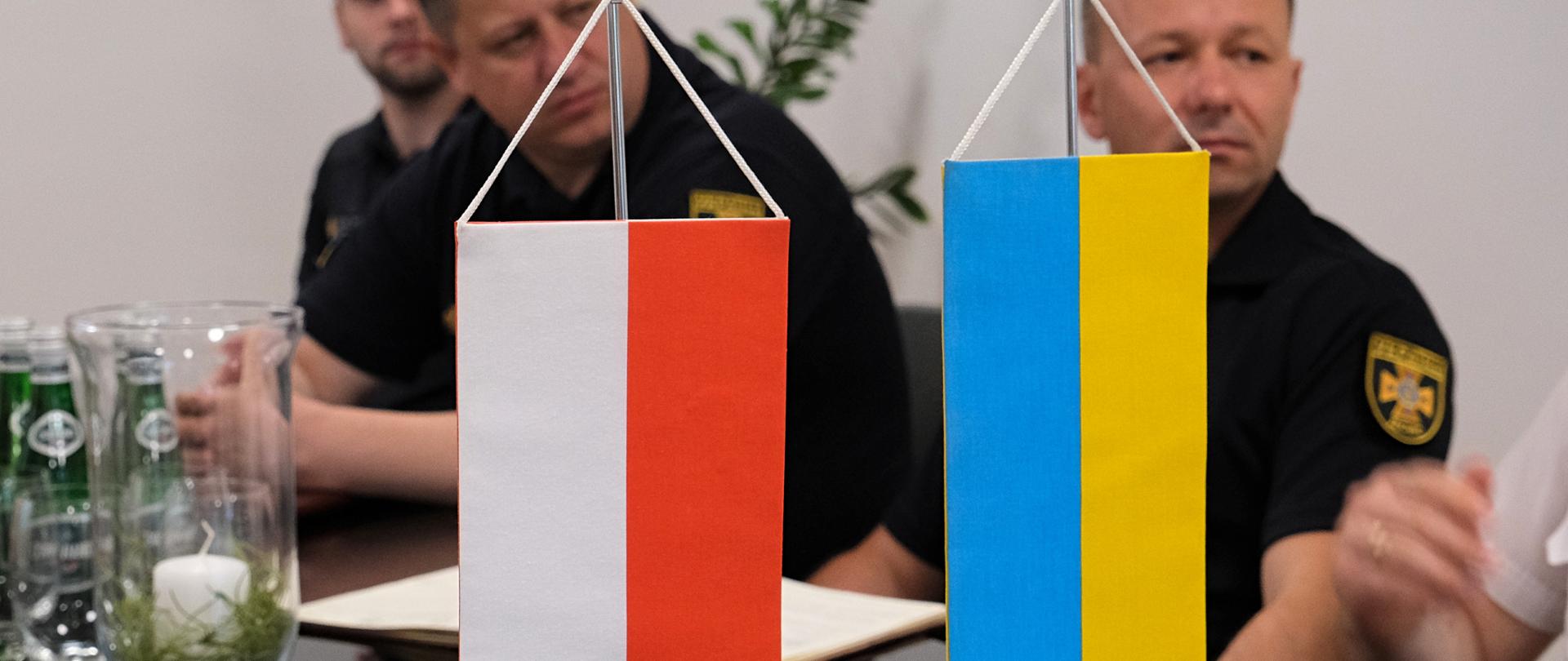 Proporczyki - flaga Polski oraz flaga Ukrainy stoją na stole, za nimi w tle 3 ukraińskich strażaków