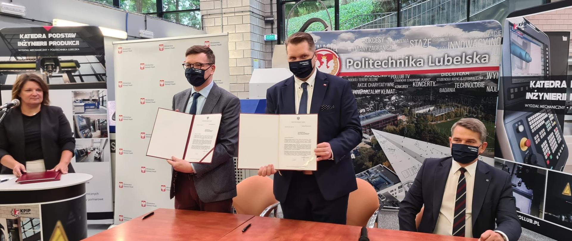 Minister Przemysław Czarnek i drugi mężczyzna stoją przy stole i trzymają w ręku teczki z podpisanym dokumentem. Na twarzy mają maseczki. Po prawej stronie ministra Czarnka mężczyzna w maseczce siedzi przy stole. Po lewej stronie kobieta przy mównicy i mikrofonie. W tle baner z panoramą Politechniki Lubelskiej. Za banerem duże okno, przez które widać drzewa. 