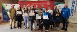 Grupa około 20 dzieci i młodzieży stoi w przestronnej sali konferencyjnej. Część z nich trzyma w rękach pamiątkowe dyplomy i białe torby z upominkami. Wraz z nimi pozują strażacy w piaskowych mundurach służbowych oraz mężczyźni w garniturach i niebieskiej kurtce. W tle widoczna jest duża czerwona ścianka z godłem Polski i napisem „Państwowa Straż Pożarna – Komenda Powiatowa w Skarżysku-Kamiennej” oraz baner reklamowy Castorama.
