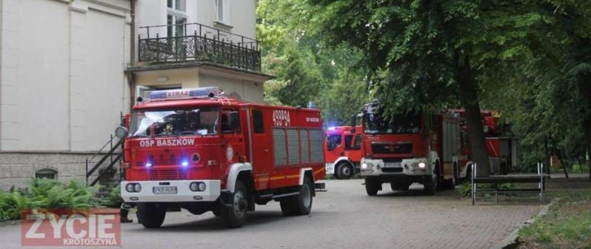 Trzy pojazdy gaśnicze zaparkowane przy budynku domu pomocy społecznej. Pojazdy mają włączone sygnały świetlne co świadczy o podjęciu interwencji przez strażaków w obiekcie. W tle widać park na terenie placówki.