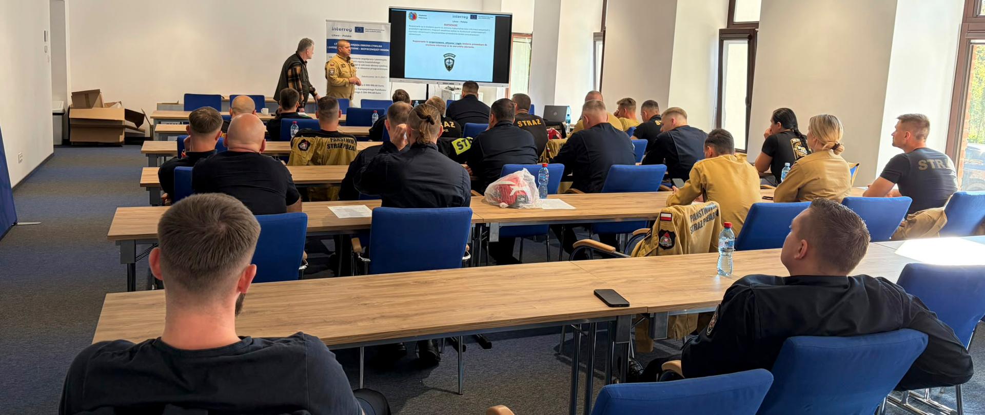 Trwa polsko-litewskie szkolenie grup poszukiwawczo-ratowniczych w ramach projektu LTPL00351 - sala wykładowa, w tle 2 wykładowców, uczestnicy przy stołach tyłem do aparatu