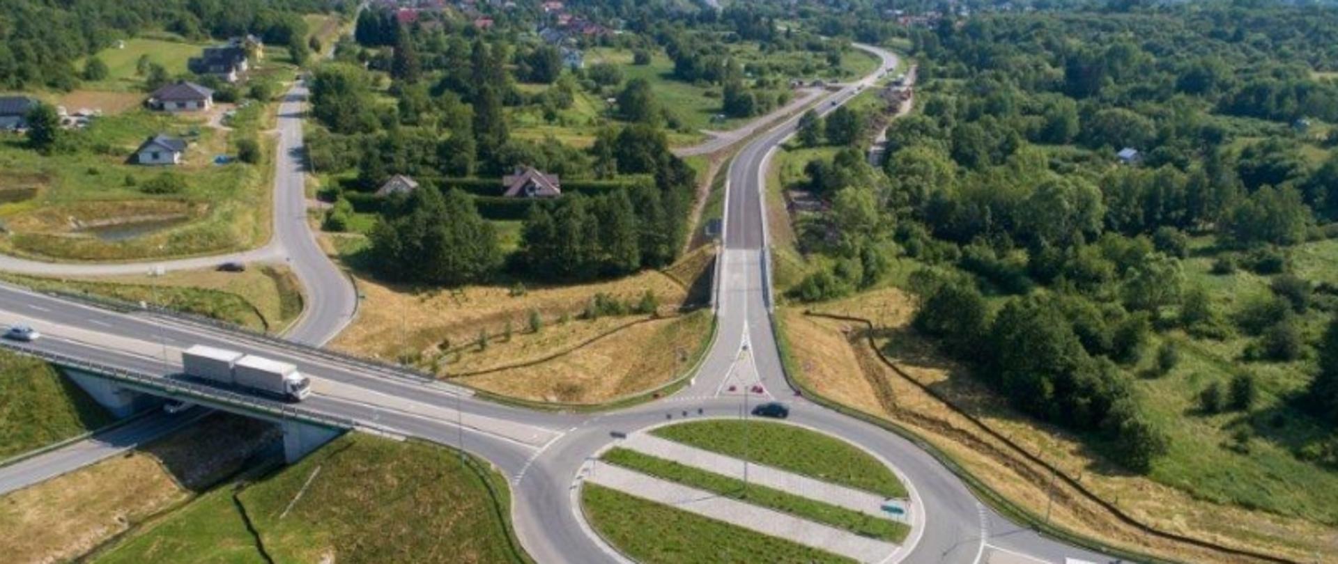 Aerial view of a roundabout with multiple connecting roads and a bridge with a truck on it, surrounded by green fields, trees, and scattered houses.