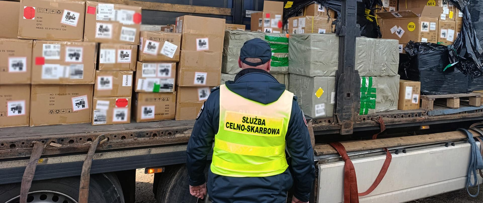 Funkcjonariusz Służby Celno‑Skarbowej stoi przy otwartej naczepie ciężarówki wypełnionej kartonami z towarem.