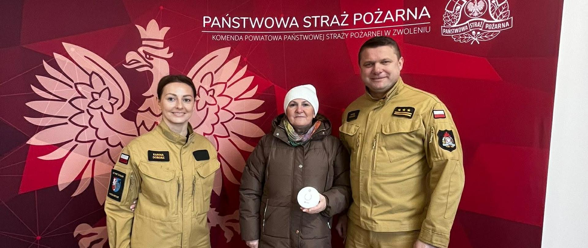 Trzy osoby pozują do zdjęcia przed ścianką Państwowej Straży Pożarnej. Pośrodku mężczyzna trzyma czujnik dymu, po jego bokach stoją dwaj strażacy w umundurowaniu służbowym.