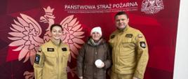 Trzy osoby pozują do zdjęcia przed ścianką Państwowej Straży Pożarnej. Pośrodku mężczyzna trzyma czujnik dymu, po jego bokach stoją dwaj strażacy w umundurowaniu służbowym.