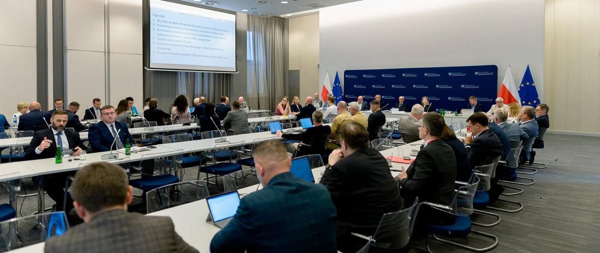 grupa oficjeli siedzi przy stole konferencyjnym, w tle wyświetlana jest prezentacja