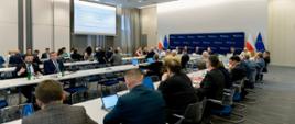 grupa oficjeli siedzi przy stole konferencyjnym, w tle wyświetlana jest prezentacja