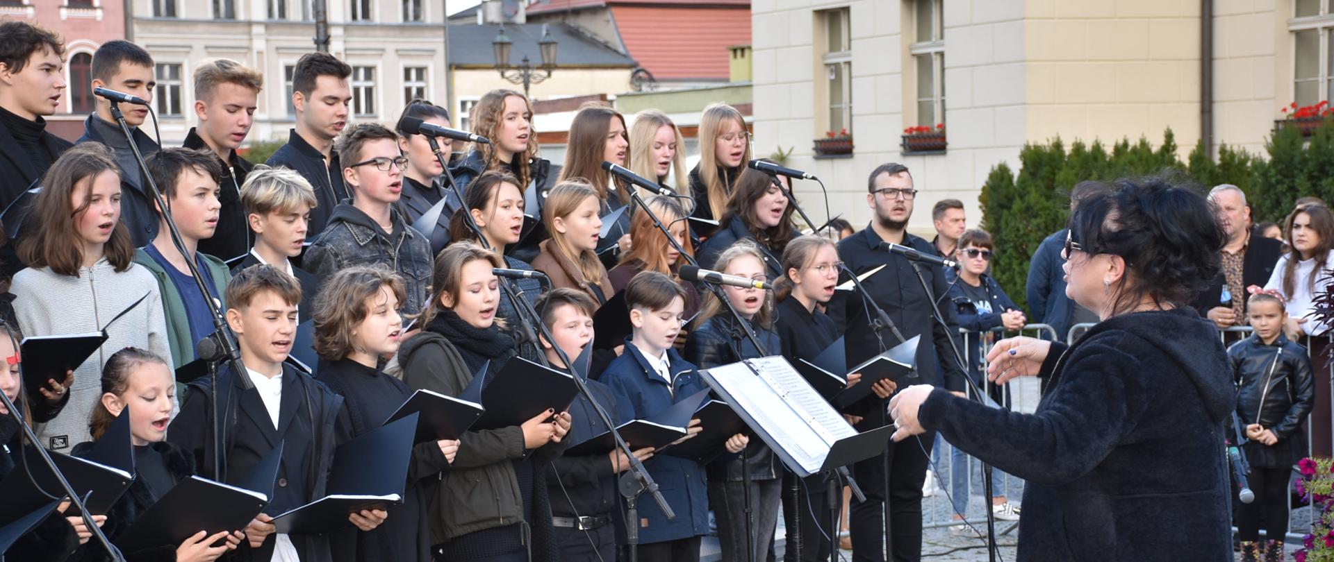 Zdjęcie przedstawia chór w trakcie występu. Członkowie chóru ubrani na czarno stoją od lewej strony pod lekkim skosem w trzech rzędach na montowanych schodach. W dłoniach trzymają otwarte czarne teczki z nutami. Przed chórem ustawione są mikrofony. Przed mikrofonami twarzą do chórzystów stoi ubrania na czarno pani dyrygent z uniesionymi rękoma (w trakcie dyrygowania). Przed sobą ma pulpit na statywie, a na nim otwartą teczkę z nutami. W tle chóru znajdują się stare kamienice w różnych kolorach (pudrowy róż, beż). Z prawej strony, w tle dyrygenta widoczni są zgromadzeni ludzie, stojący i oglądający występ chóru. 