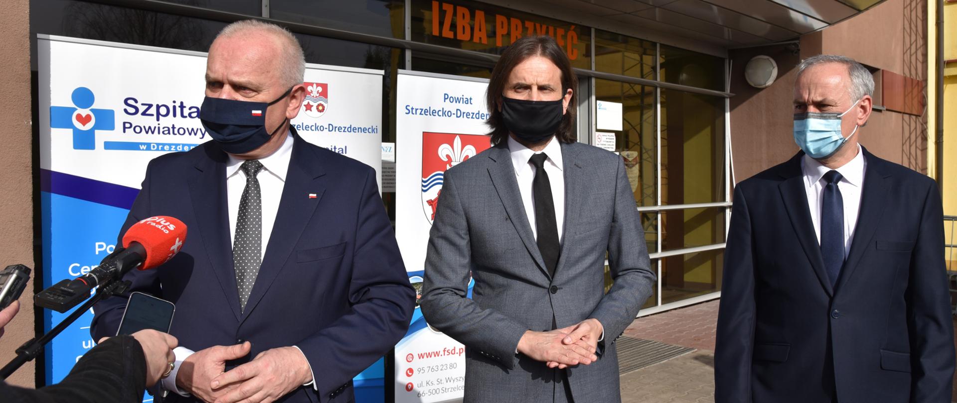 Wojewoda odpowiada na pytania dziennikarzy, obok niego stoi dyrektor lubuskiego oddziału Narodowego Funduszu Zdrowia Piotr Bromber i starosta Strzelecko-drezdenecki. W tle wejście na Izbę Przyjęć szpitala w Drezdenku oraz dwa roll-upy reklamowe szpitala